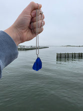 Load image into Gallery viewer, A hand holding a silver chain necklace with a bright blue teardrop-shaped pendant, set against a background of calm water and a wooden pier.