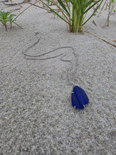 Load image into Gallery viewer, A silver-toned chain necklace with a dark blue teardrop-shaped pendant lies on a textured gray concrete surface next to some green grass.