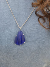 Load image into Gallery viewer, A silver chain necklace with a carved blue glass pendant, displayed on a textured blue-gray knit fabric next to a strand of brown hair.