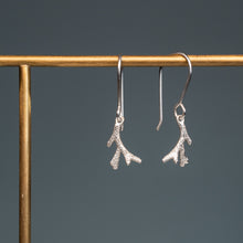 Load image into Gallery viewer, A pair of silver drop earrings shaped like textured tree branches or small antlers, hanging from a gold-tone display stand against a dark gray background.