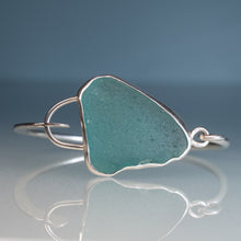 Load image into Gallery viewer, A sterling silver cuff bracelet featuring a large, irregularly shaped sea glass cabochon in a dusty teal green color, displayed on a reflective white surface against a soft blue-gray gradient background.