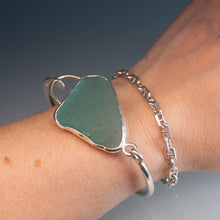Load image into Gallery viewer, A sterling silver cuff bracelet featuring a large, irregularly shaped green sea glass inlay, shown worn on a person's wrist alongside a silver chain bracelet.
