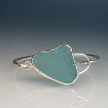 Load image into Gallery viewer, A sterling silver open cuff ring featuring a rough, organic-shaped sea glass cabochon in a muted teal green color, displayed on a reflective surface against a dark gradient background.