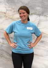 Load image into Gallery viewer, Woman wearing a blue t-shirt with a logo against a marble wall.