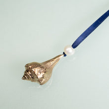 Load image into Gallery viewer, close up back of cast bronze lightning whelk ornament from key west florida with freshwater pearl and navy blue satin ribbon by hkm jewelry on blue background