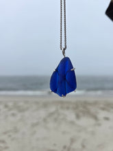 Load image into Gallery viewer, A blue glass seashell pendant on a silver chain, photographed against an out-of-focus beach background.

