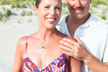 Load image into Gallery viewer, A smiling couple standing on a sandy beach with dune grass in the background. The woman wears a colorful patterned camisole and a heart-shaped pendant necklace, while the man wears a white polo shirt and holds her hand, which features a large ring.
