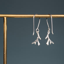 Load image into Gallery viewer, A pair of silver drop earrings shaped like small coral branches, hanging from a gold-tone display stand against a dark slate blue background.

