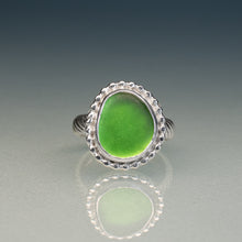 Load image into Gallery viewer, A silver ring featuring a large, irregularly shaped green glass cabochon stone, set in a scalloped silver bezel with a twisted silver band, shown on a reflective gray surface.
