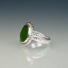 Load image into Gallery viewer, A silver ring featuring a large, oval-shaped green stone in a beaded halo setting, with a twisted cable-style band, shown on a reflective gray surface.
