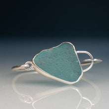 Load image into Gallery viewer, A sterling silver cuff ring featuring a large, irregularly shaped piece of textured teal-green sea glass, displayed on a reflective gray surface against a dark blue gradient background.

