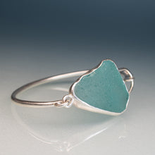 Load image into Gallery viewer, A sterling silver ring featuring an irregular, heart-shaped sea glass cabochon in a light teal blue-green color, set against a dark blue-gray gradient background.
