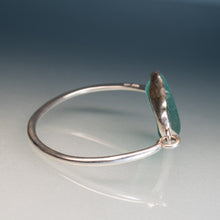 Load image into Gallery viewer, A silver-toned open bangle ring featuring a polished, irregularly shaped green stone set in a textured bezel, displayed on a reflective light gray surface against a dark blue gradient background.
