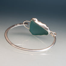 Load image into Gallery viewer, A sterling silver bangle bracelet featuring a polished, irregularly shaped green sea glass centerpiece, displayed on a reflective gray surface against a dark gradient background.
