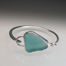 Load image into Gallery viewer, A silver-toned open bangle bracelet featuring a large, irregular-shaped sea glass charm in a muted teal green color, displayed on a neutral gray reflective surface.
