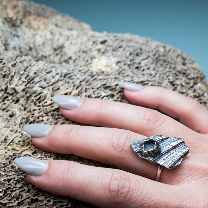 cast darkened metal textural ring with barnacle on hand model with textured beige coral background