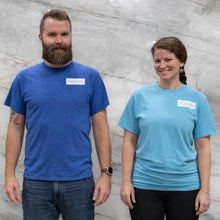Load image into Gallery viewer, Two people wearing blue t-shirts with logos against a gray textured wall.
