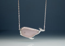 Load image into Gallery viewer, A whale-shaped pendant necklace featuring a smooth, pale pink stone inlay set in polished silver, suspended from a delicate silver chain against a dark blue gradient background.
