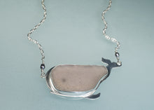Load image into Gallery viewer, A whale-shaped pendant necklace made of polished silver with a smooth, matte taupe-gray stone inlay, shown on a light blue background.

