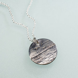 sedge island workshop marsh necklace with shorebirds in sterling silver by hkm jewelry laying on blue background