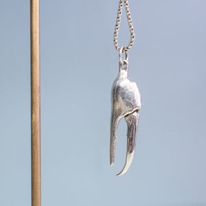 close up view of the cast fiddler crab claw necklace in recycled silver hanging from brass and coral necklace display on a blue background by hkm jewelry