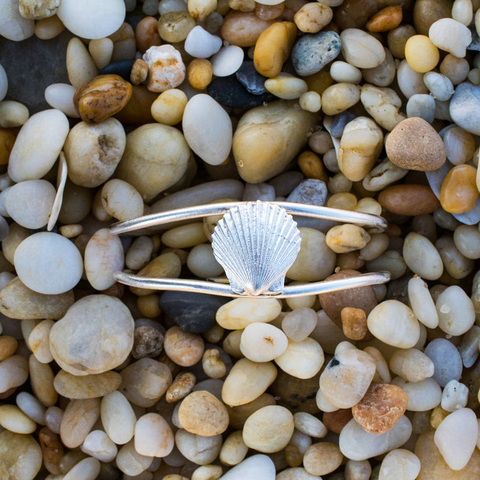 cast silver bay scallop shell cuff laying in beach pebbles by hkm jewelry hali maclaren